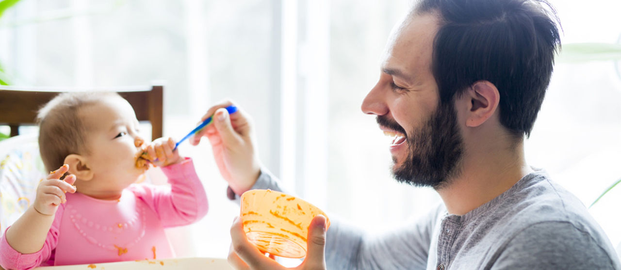 A man feeds solid foods to a baby sitting in a highchair.
